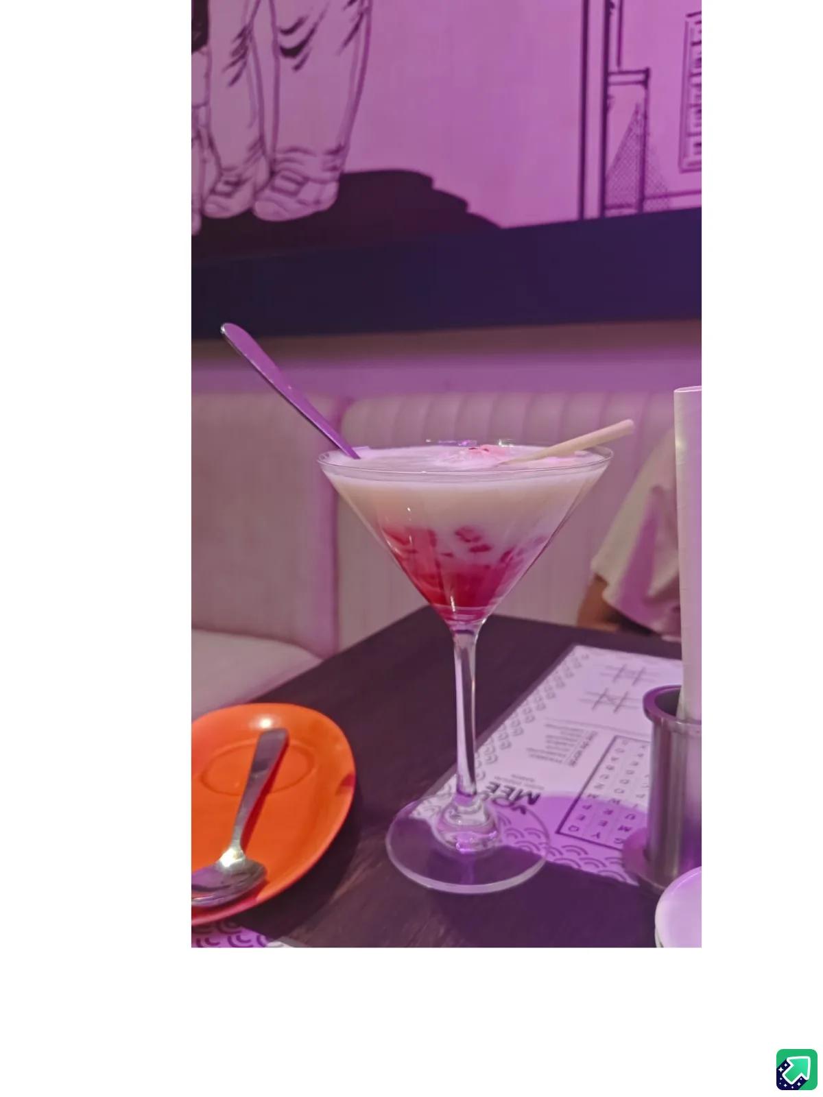 A martini glass containing a layered pink and white drink with two spoons inserted, sits on a dark table in a restaurant…