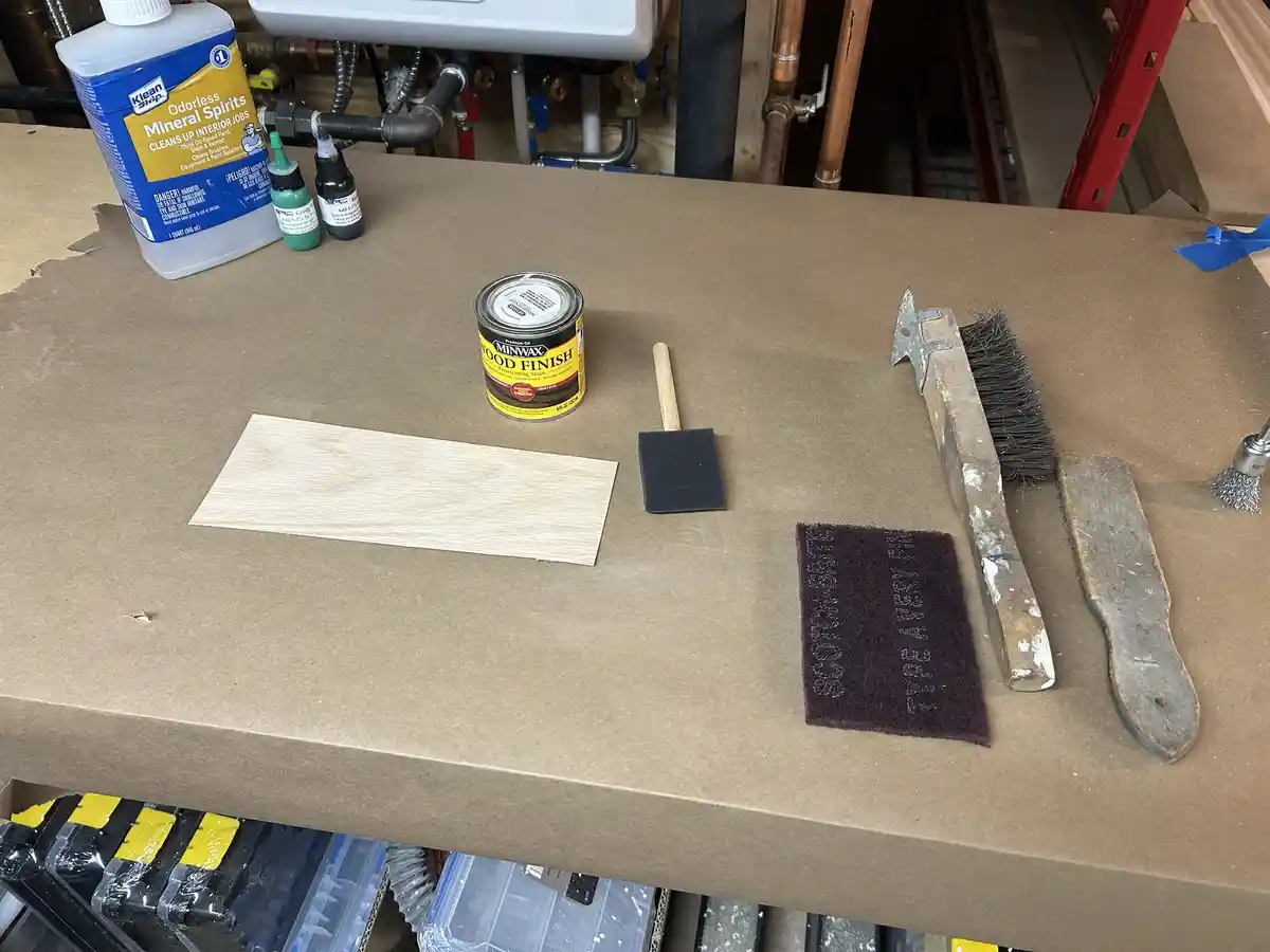 A workbench displays woodworking tools and supplies including a can of Minwax Wood Finish, a wooden plank, a foam brush, a…