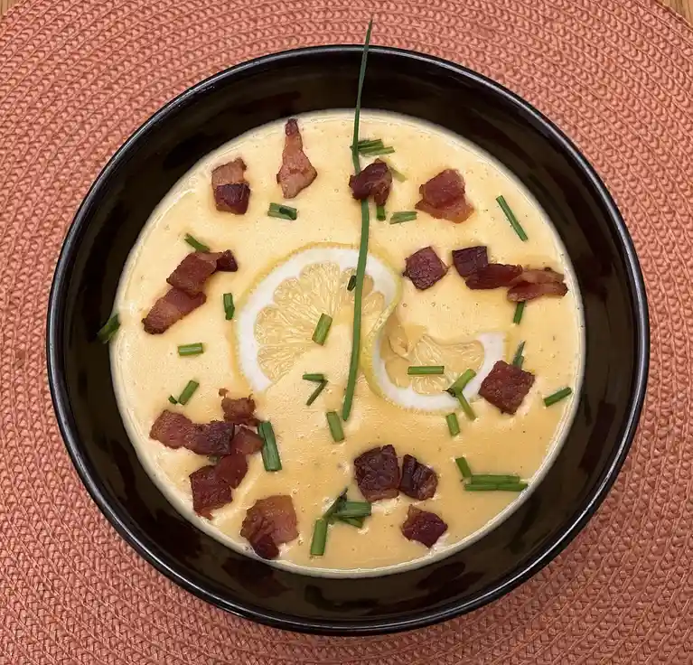 A top-down photo of a creamy yellow soup in a black bowl, garnished with crispy bacon bits, chopped green chives, and a…