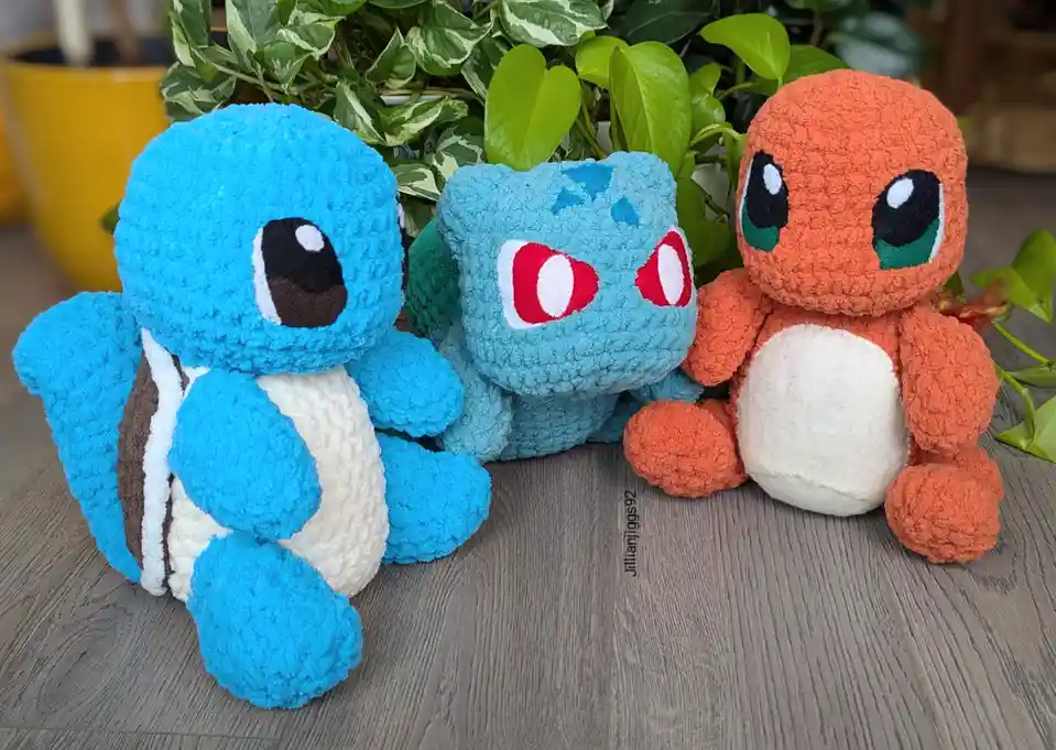 Three crocheted Pokémon plushies—Squirtle, Bulbasaur, and Charmander—are sitting side-by-side on a wooden floor in front of…
