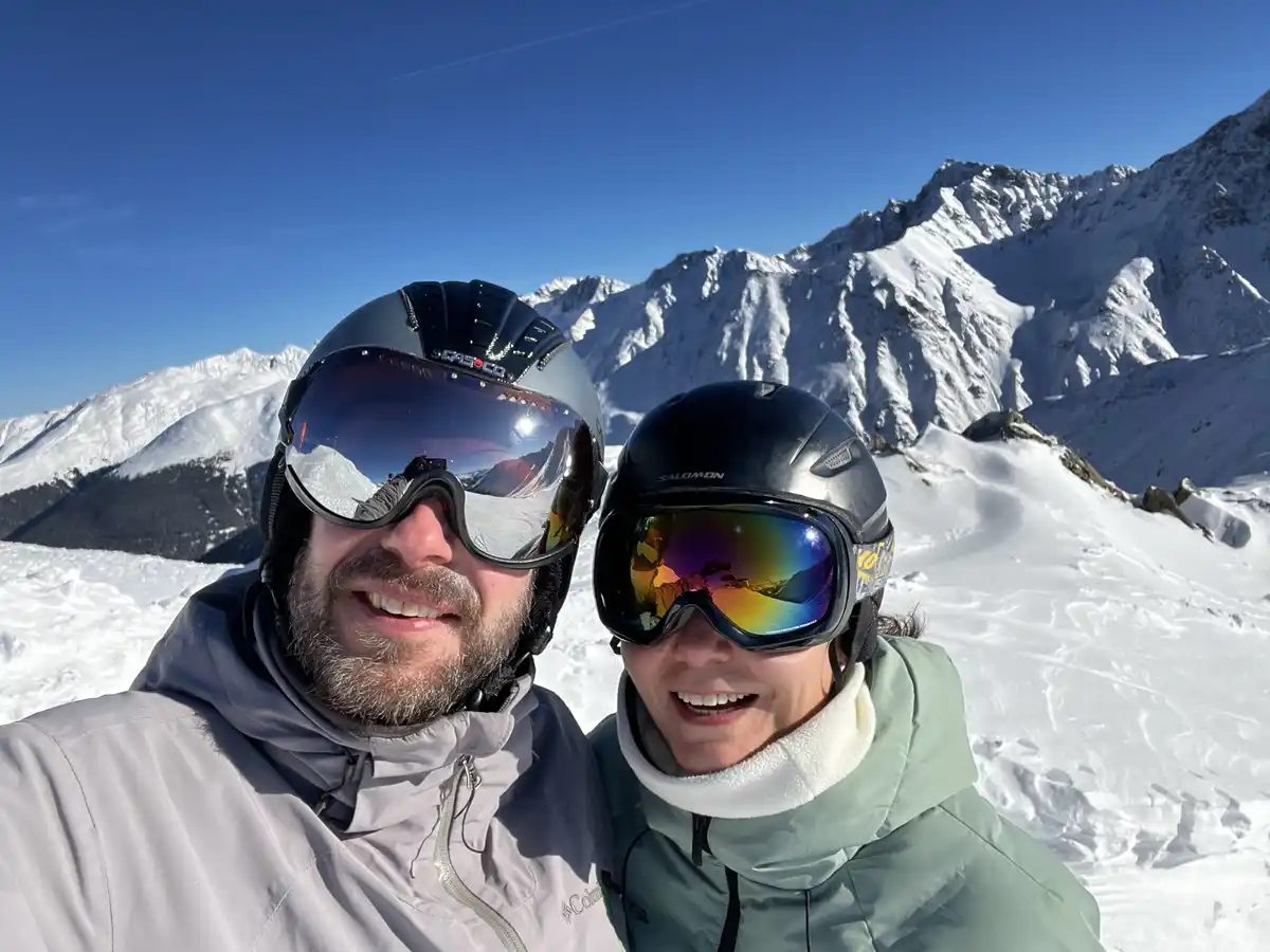 Two people in ski gear, including helmets and goggles, smile for a selfie on a snowy mountain with peaks in the background…