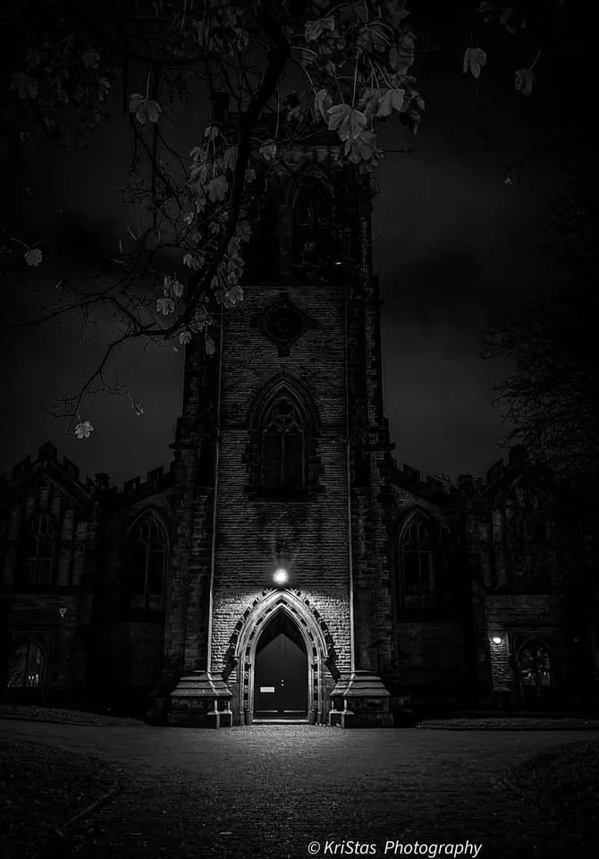 A black-and-white photo of a Gothic church at night, with a single spotlight illuminating the arched entrance. Overlaid text…