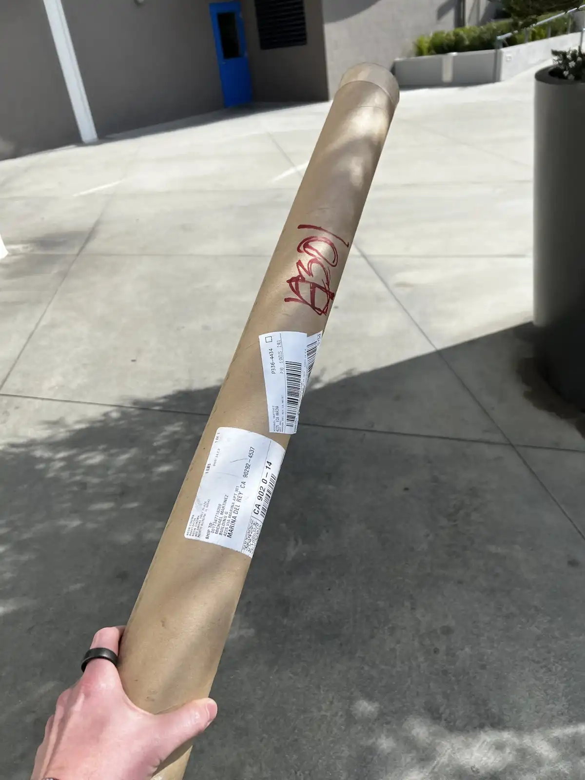 A hand holds a large, brown paper-wrapped cylindrical package outdoors on a concrete patio. The package has shipping labels…