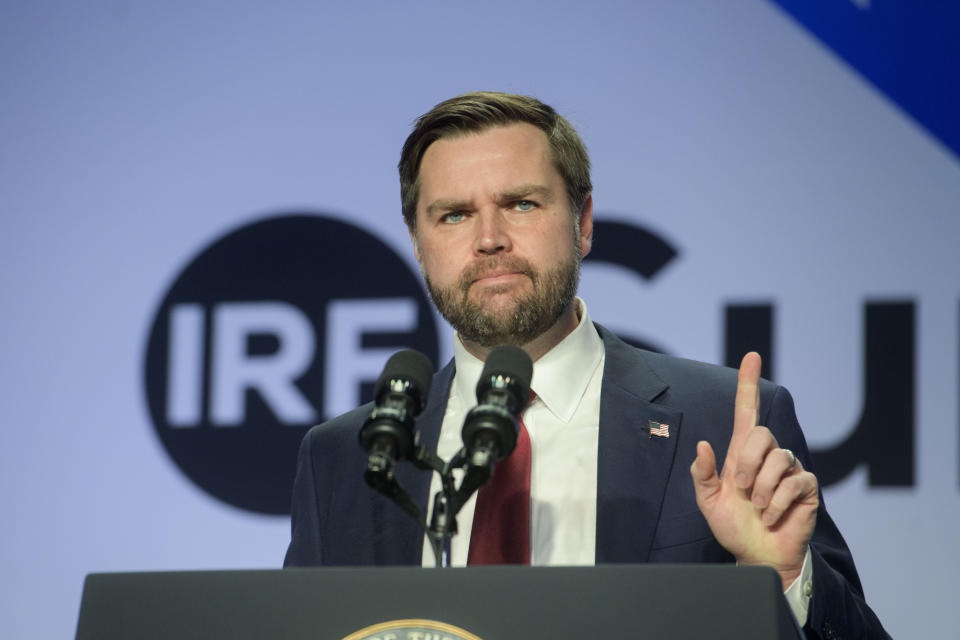 a man with a beard and suit stands at a podium with microphones, pointing his index finger upward. Behind him, a large blue…