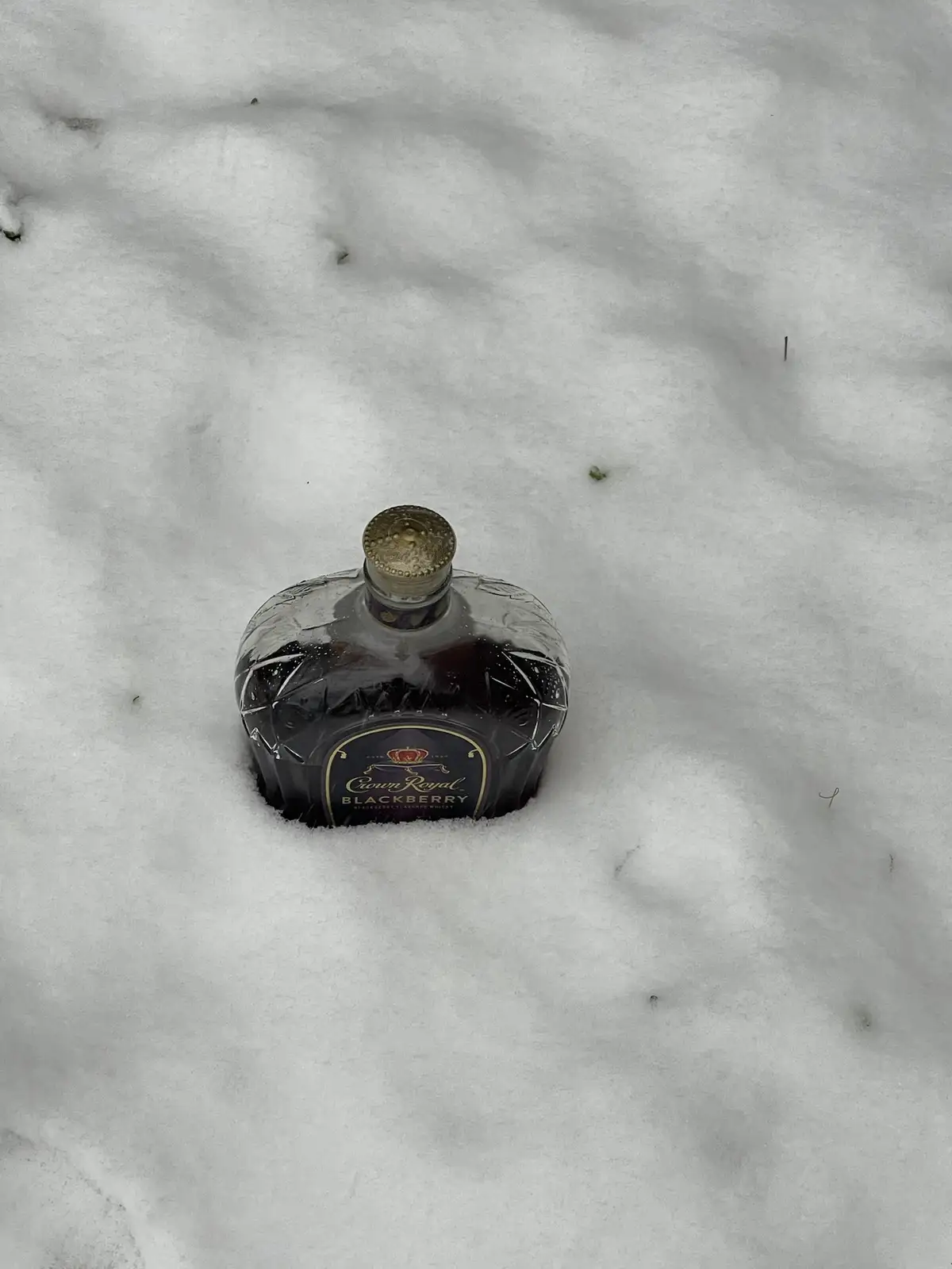A bottle of Crown Royal Blackberry whisky is partially buried in snow, with only the top and label visible. The bottle has a…