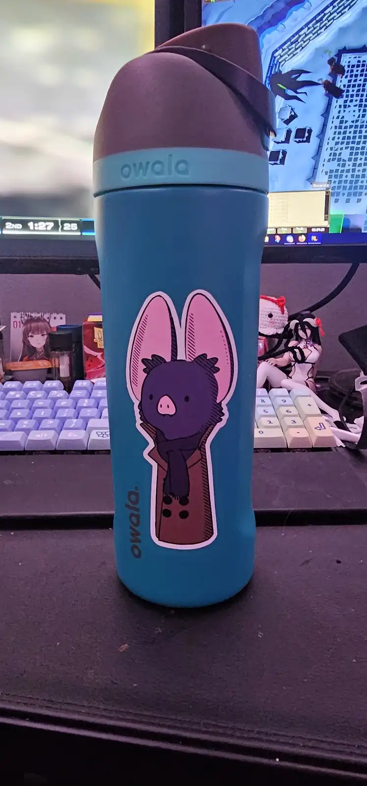 A teal Owala water bottle with a cartoon bat sticker sits on a desk in front of a computer monitor displaying a game. The…