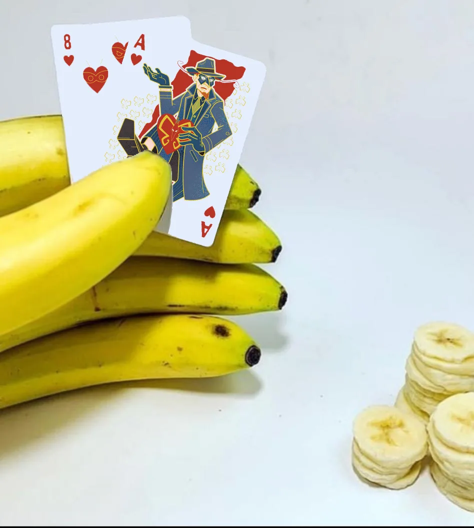 Two playing cards, the 8 of hearts and Ace of hearts, are propped on top of a bunch of whole bananas, with stacks of sliced…