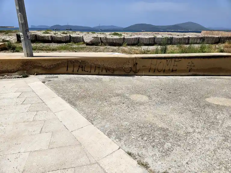 A low concrete wall near a seaside path has the words 'ITALIANS GO HOME' spray-painted in black graffiti. Behind the wall is…