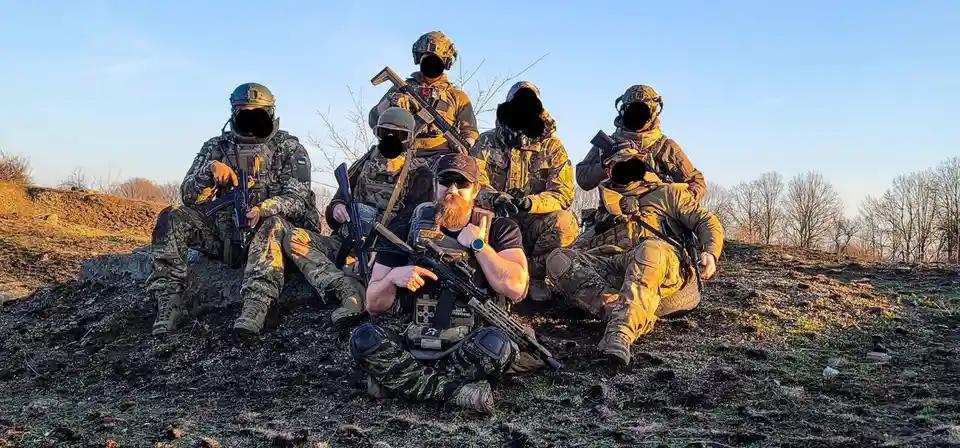 A group of six men in full tactical gear, including helmets, camouflage uniforms, and rifles, are posing for a photo…