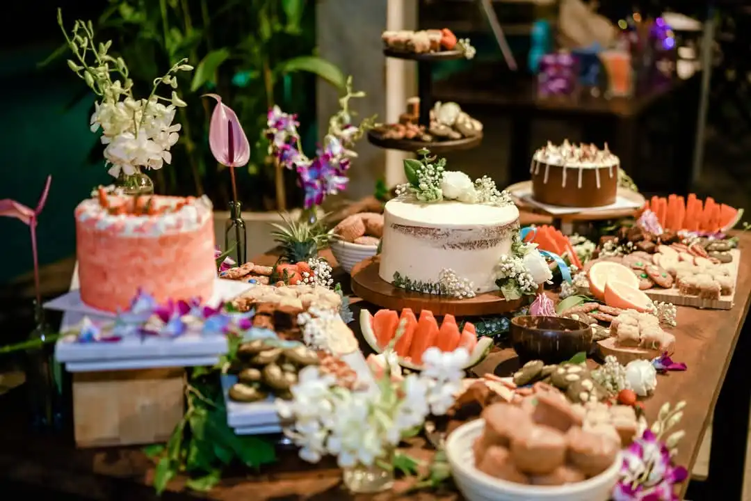 A lavish dessert table features multiple cakes, watermelon slices, cookies, and assorted sweets, all adorned with white and…