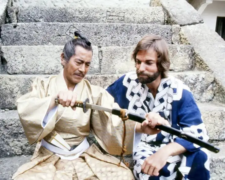 Two men sit on stone steps, one in a gold kimono holding a katana, the other in a blue and white patterned kimono holding…