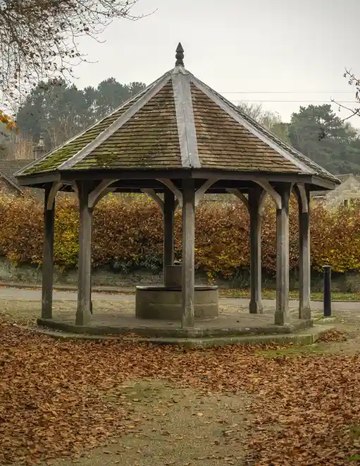 An outdoor stone well covered by a wooden gazebo with a mossy roof, surrounded by fallen autumn leaves and hedge. The…