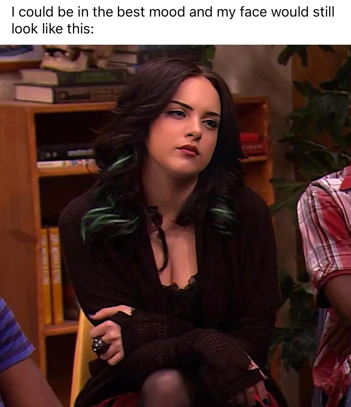 A woman with dark hair and a black cardigan sits with arms crossed, looking unimpressed or bored. Behind her is a wooden…