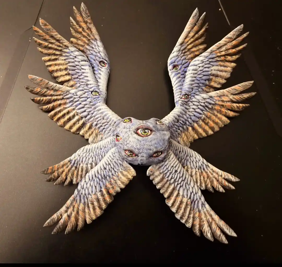 A sculpted, multi-winged bird-like creature with a blue-gray body and brown-tipped feathers, spread wide on a dark surface…