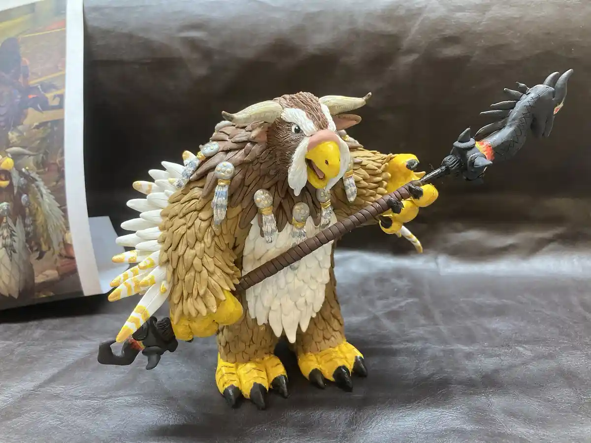 A detailed figurine of a fantasy creature resembling a bird with horns, brown and white feathers, and yellow feet, holding a…