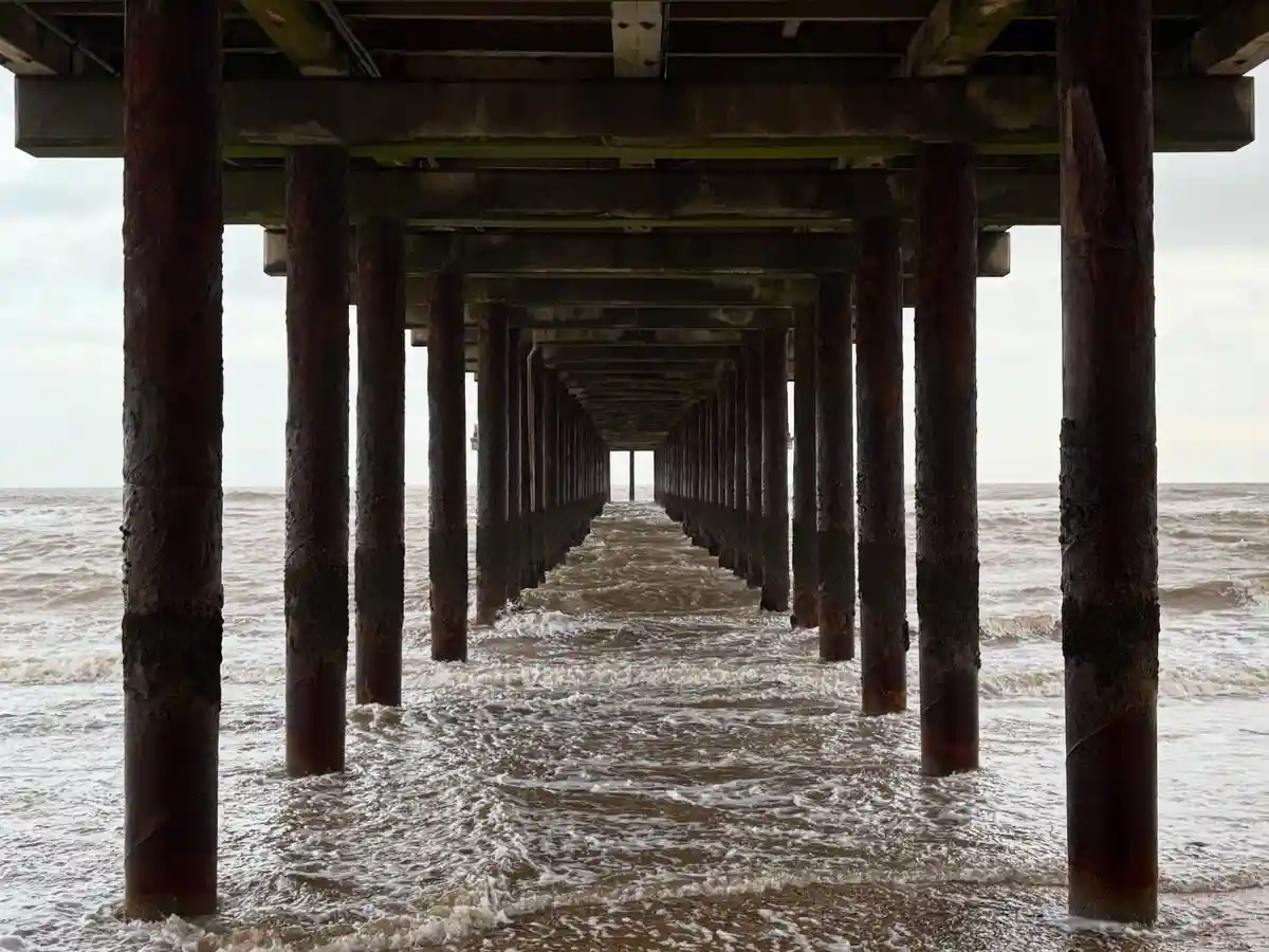 A photograph taken from under a weathered wooden pier, showing rows of rusty support pillars extending into the ocean under…