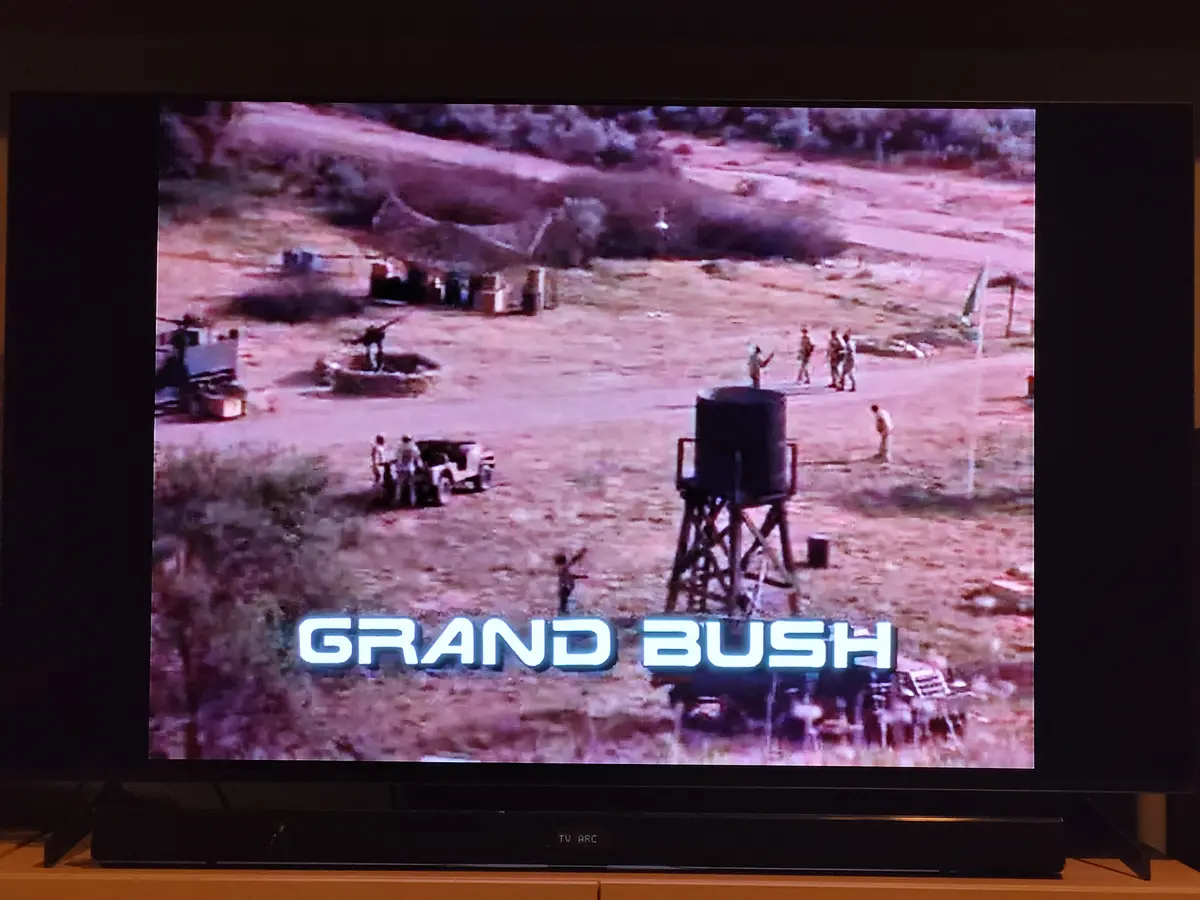 A television screen displays a grainy, reddish-tinted outdoor scene with several people and vehicles in a barren field…