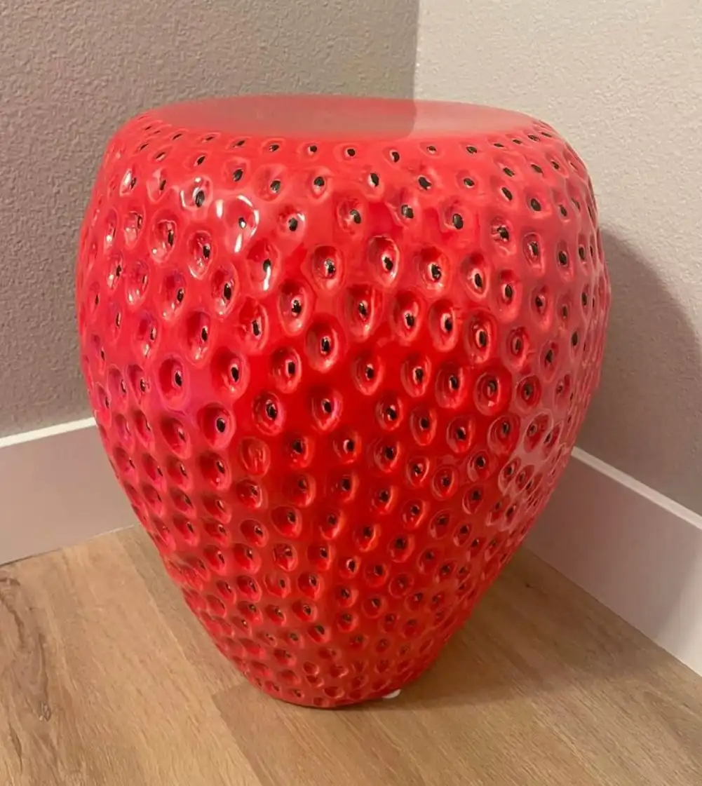A glossy, oversized red stool shaped like a strawberry with black seeds, sitting on a wooden floor against a beige wall. The…