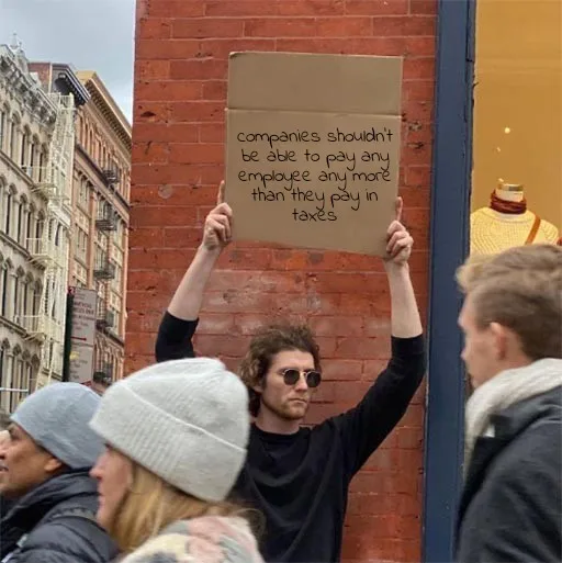 A man with curly hair and sunglasses holds up a cardboard sign with handwritten text in front of a brick wall, surrounded by…
