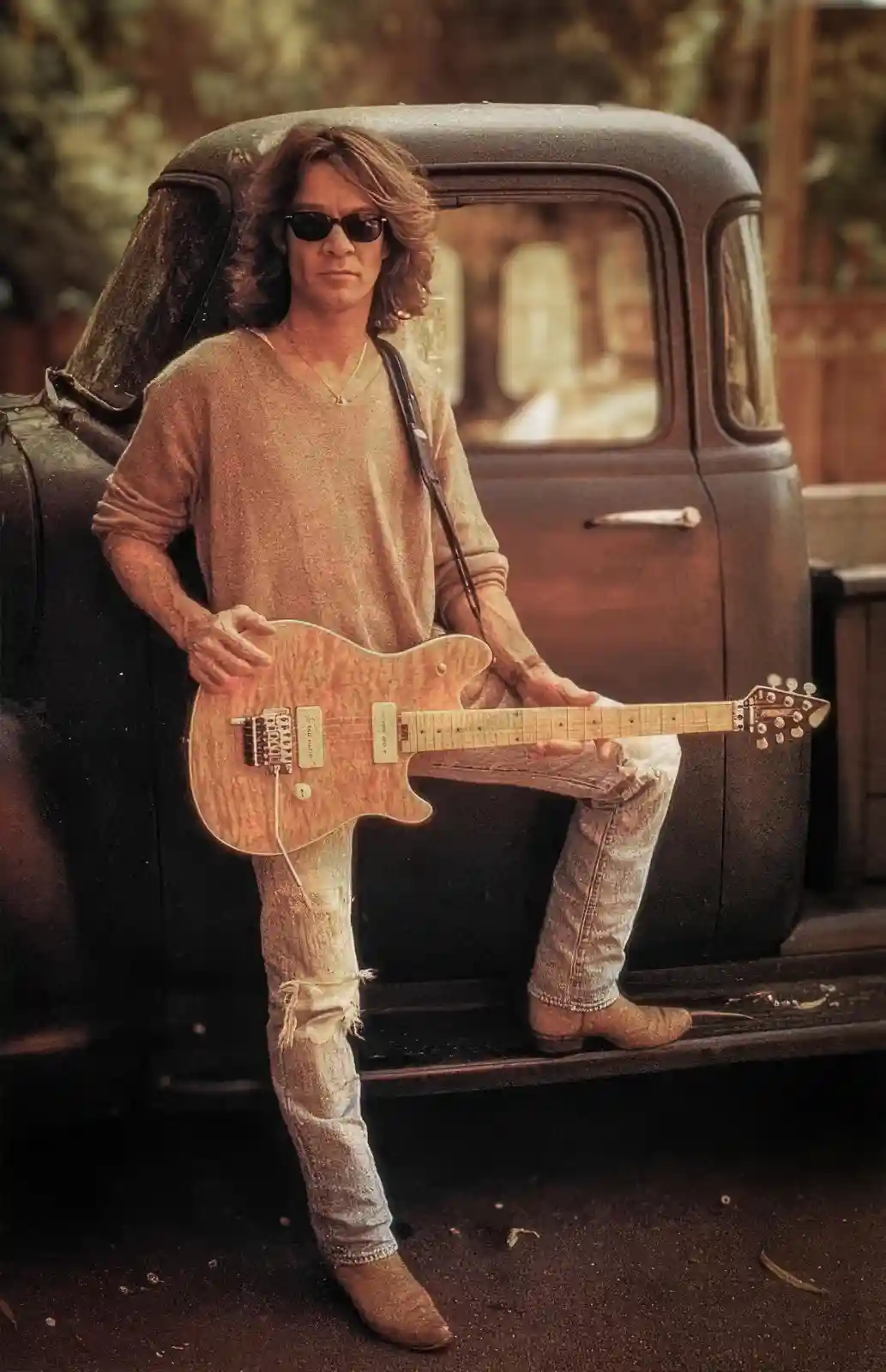A man with long hair and sunglasses sits on the tailgate of a vintage pickup truck, holding an electric guitar. He wears a…