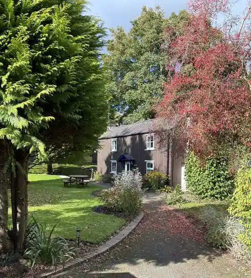 A picturesque view of a quaint brown wooden cottage surrounded by lush greenery and vibrant red foliage, with a gravel path…