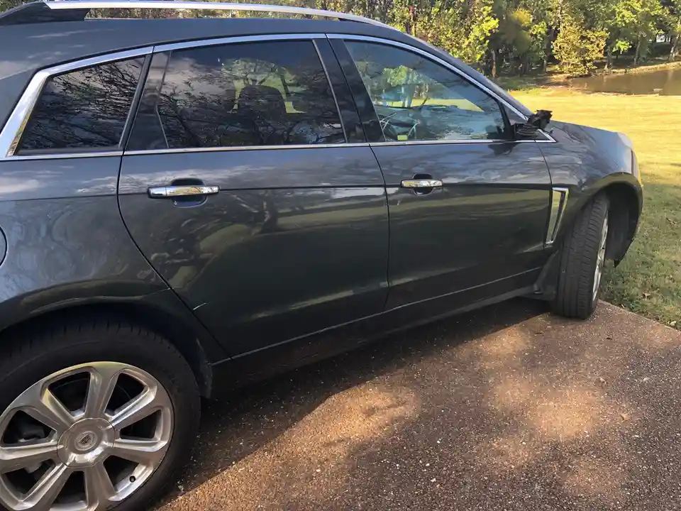 A dark gray SUV is parked on a gravel driveway with trees and a body of water in the background. The car's side profile is…