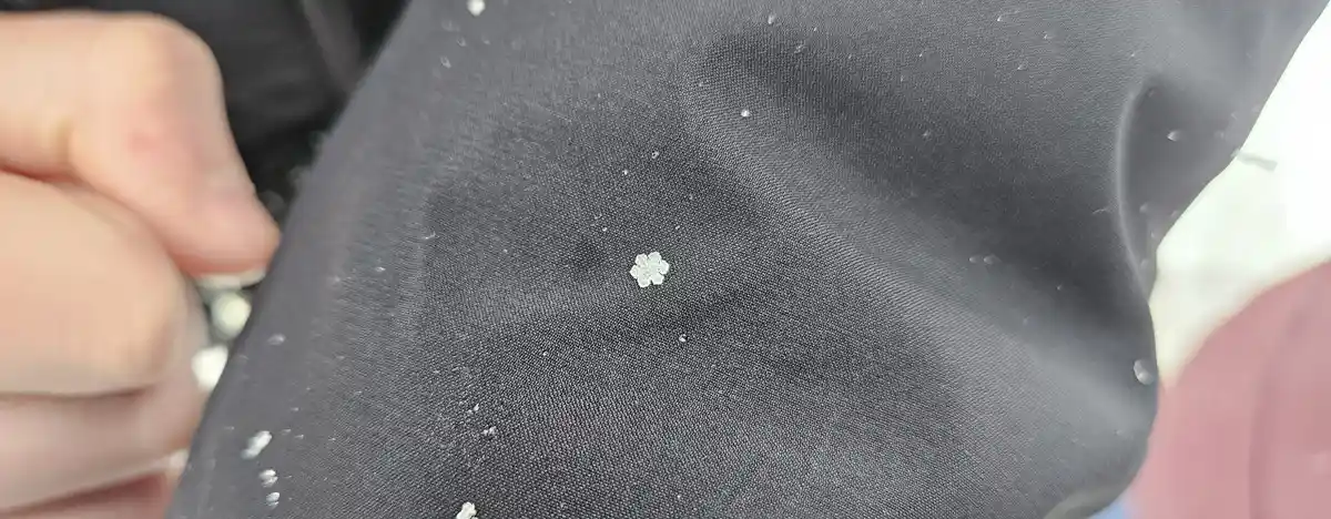 A close-up photo shows a small, intricate snowflake resting on the dark fabric of a jacket, with other tiny flakes scattered…