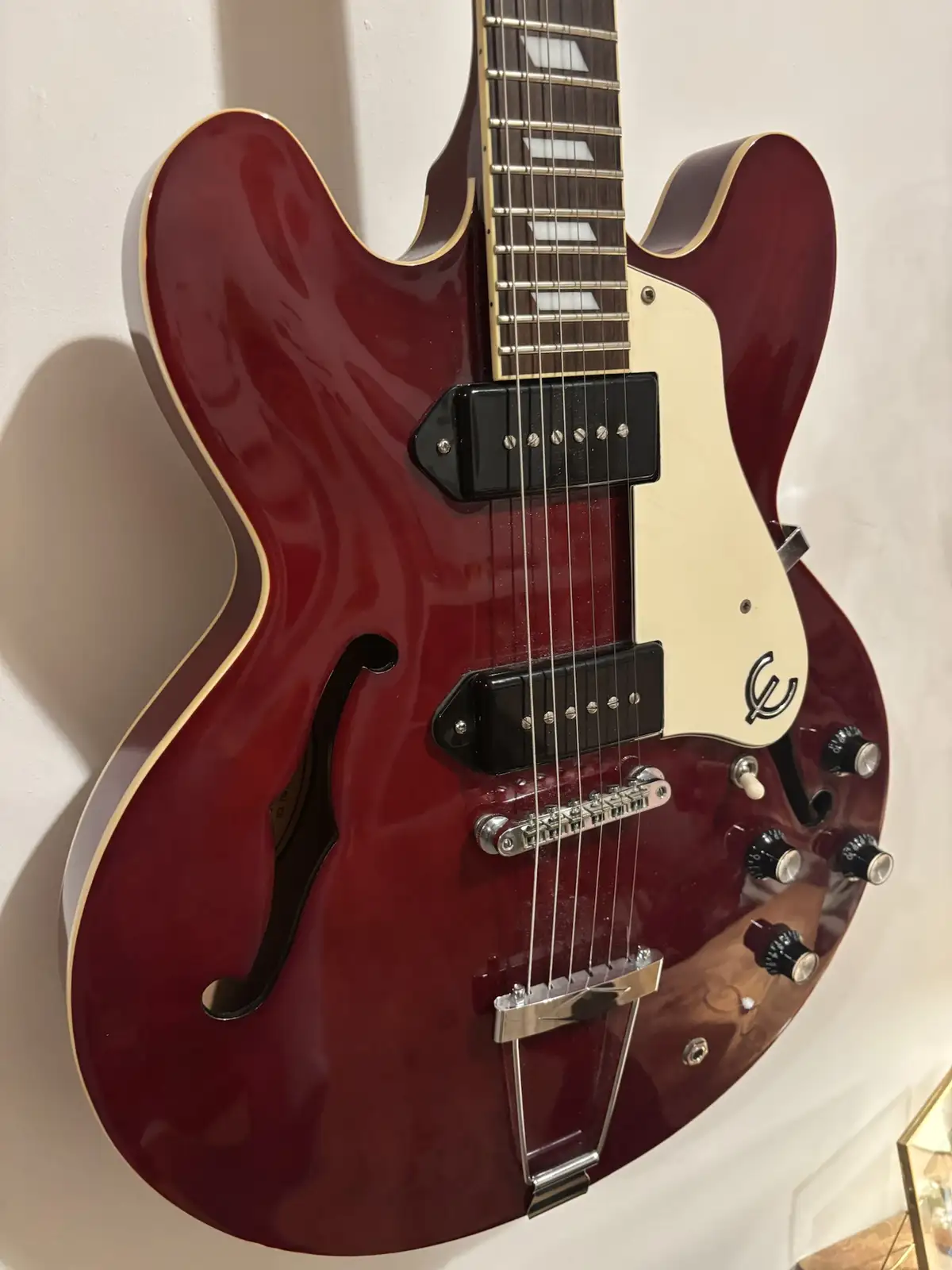 A glossy, deep red semi-hollow body electric guitar with a white pickguard and black pickups is mounted on a white wall. The…