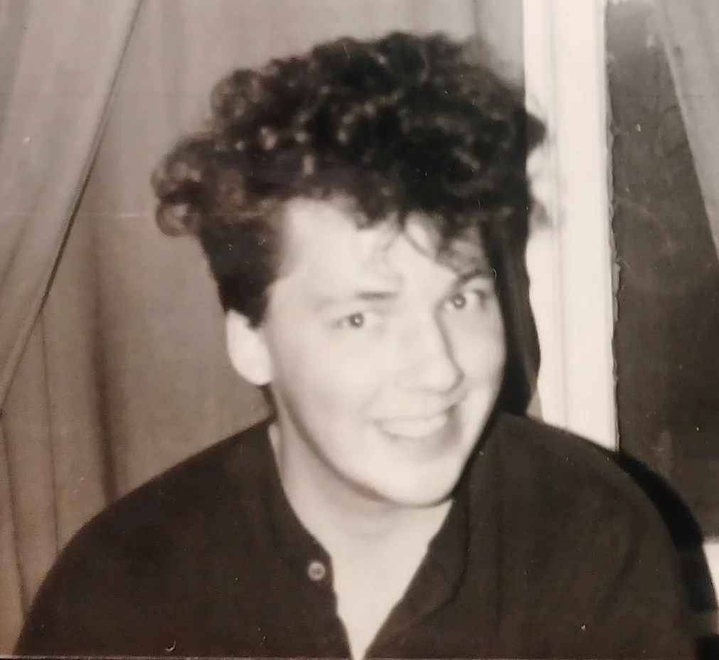 A grainy, black-and-white photo shows a young person with curly hair, smiling and looking slightly to the side, wearing a…