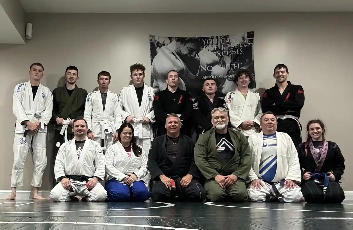 A group photo of about thirteen people in martial arts gi uniforms posing on a mat in a gym. Behind them, a large…