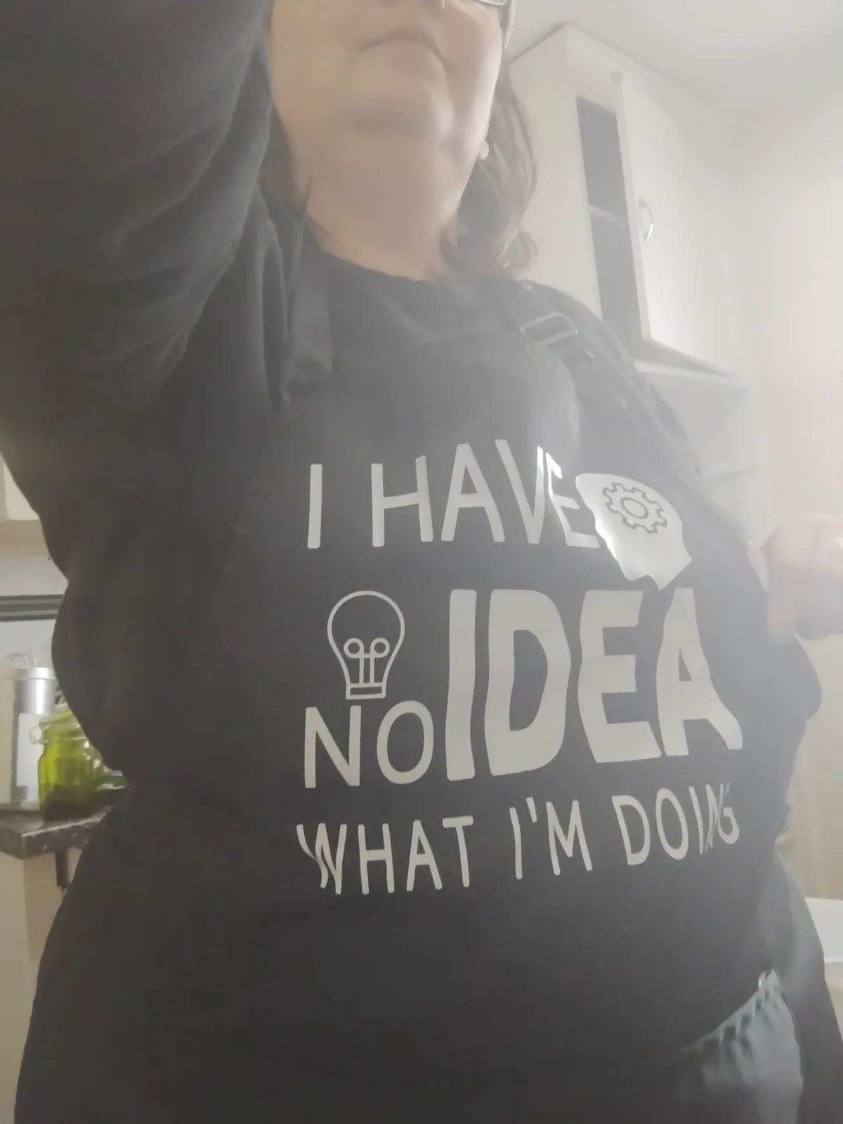 A person wearing a black apron with white text and icons that reads 'I HAVE NO IDEA WHAT I'M DOING', featuring a lightbulb…