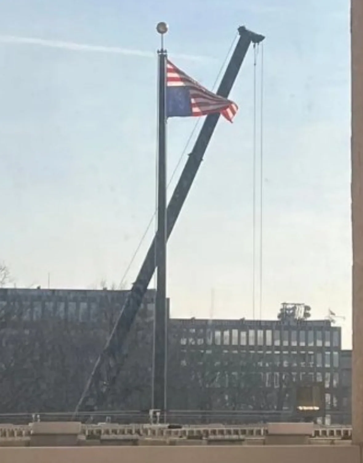 An american flag is visible on a flagpole, but a large crane boom is positioned diagonally across it, appearing to lower the…