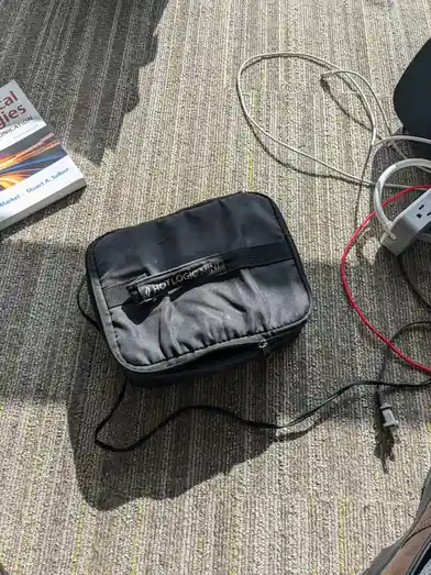 A black rectangular zippered case labeled 'HOTLOGIC MINI' sits on a striped carpet next to a textbook, tangled white cables…