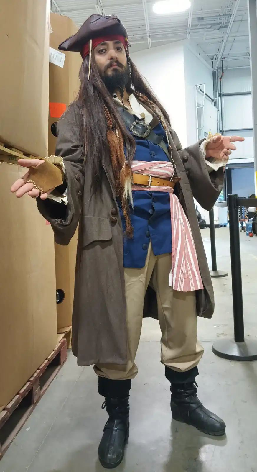 A man dressed as a pirate with a tricorn hat, long braided hair, and a beard stands in a warehouse next to large cardboard…