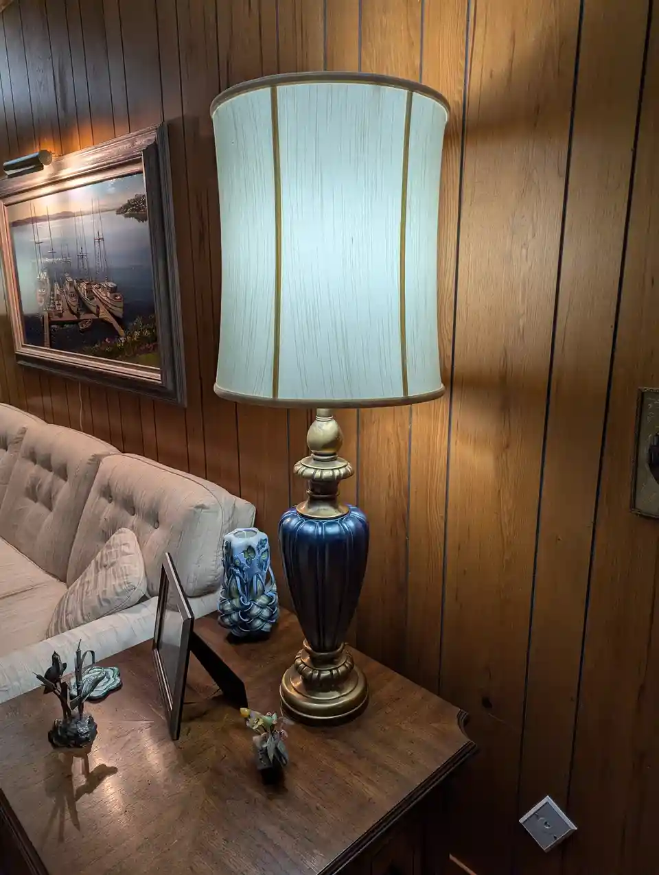 A close-up photo of a blue ceramic table lamp with a light-colored pleated shade sitting on a wooden side table next to a…