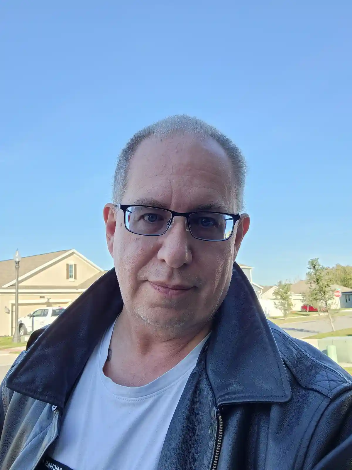 A middle-aged man with glasses and a black leather jacket stands outdoors under a clear blue sky, looking directly at the…