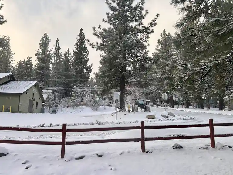A snowy outdoor scene with a red wooden fence in the foreground, snow-covered pine trees, and a building labeled…