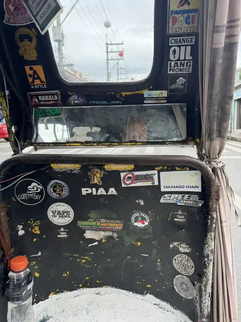 The image shows the interior of a heavily stickered vehicle, likely a Philippine tricycle, with a dirty window and a…