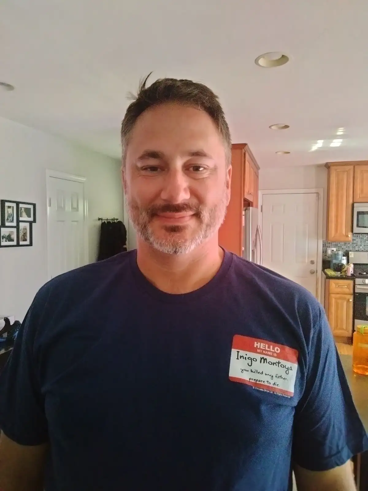 A man with a beard and short hair smiles while wearing a dark blue t-shirt with a name tag that reads 'HELLO MY NAME IS…