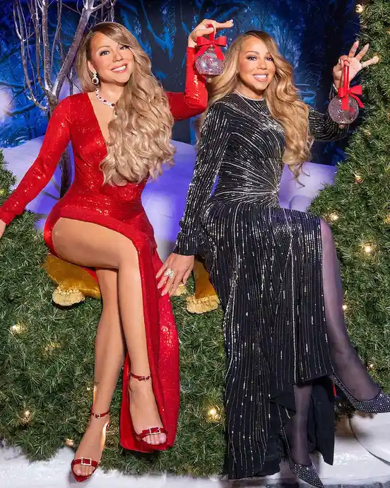Two women, Mariah Carey and Beyoncé, are posing together in glamorous, sequined dresses—one red, one black—sitting on a…