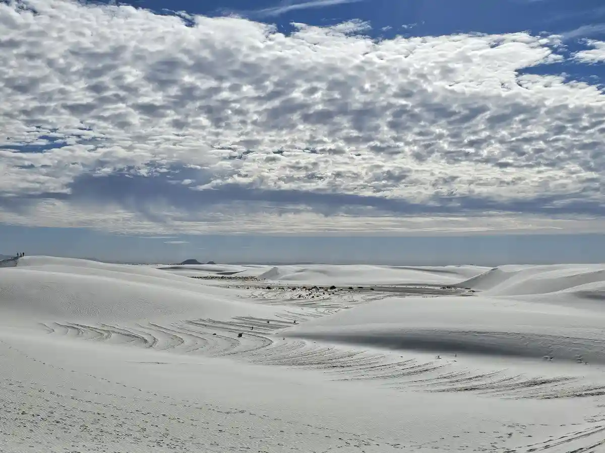 A wide landscape photo of white sand dunes under a partly cloudy blue sky, with faint tire tracks and footprints visible on…