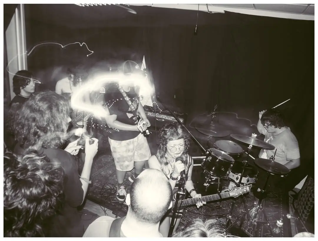 A black-and-white photo of a band performing live, with a guitarist in the center having a bright, glowing light trail…