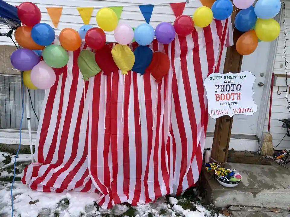 A red-and-white striped photo booth backdrop decorated with colorful balloons and a pennant banner, set outdoors in snow. A…