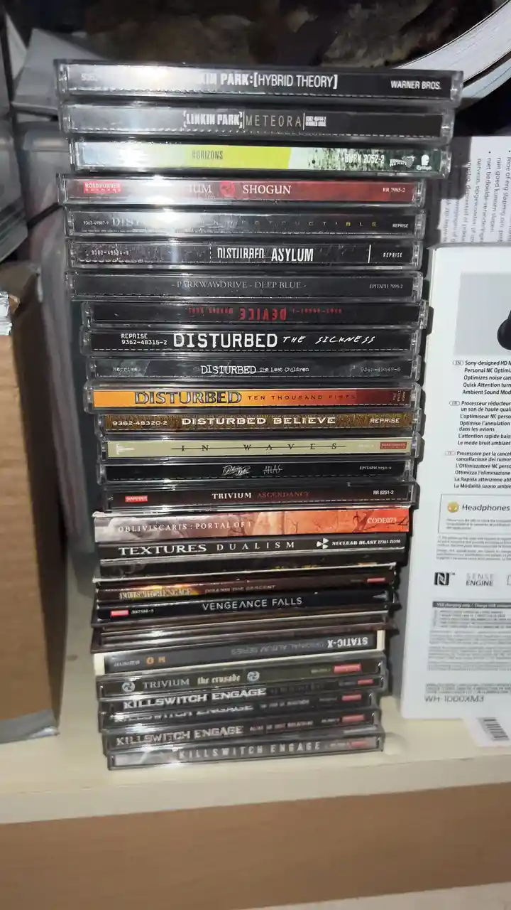 A tall stack of cassette tapes featuring metal bands like Linkin Park, Disturbed, Trivium, and Killswitch Engage, with album…