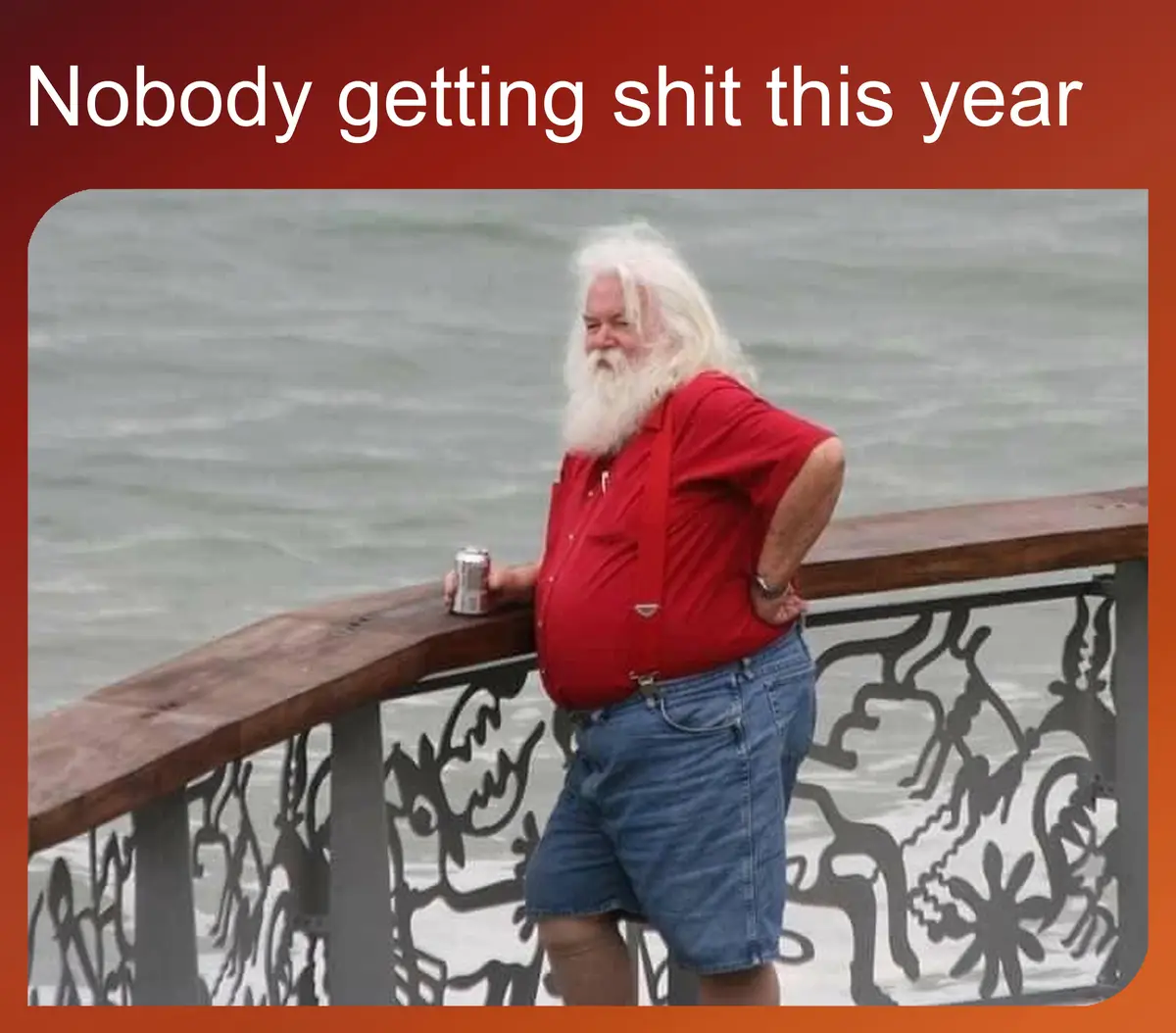 A man with long white hair and a full beard, wearing a red shirt and blue shorts, leans against a decorative railing…