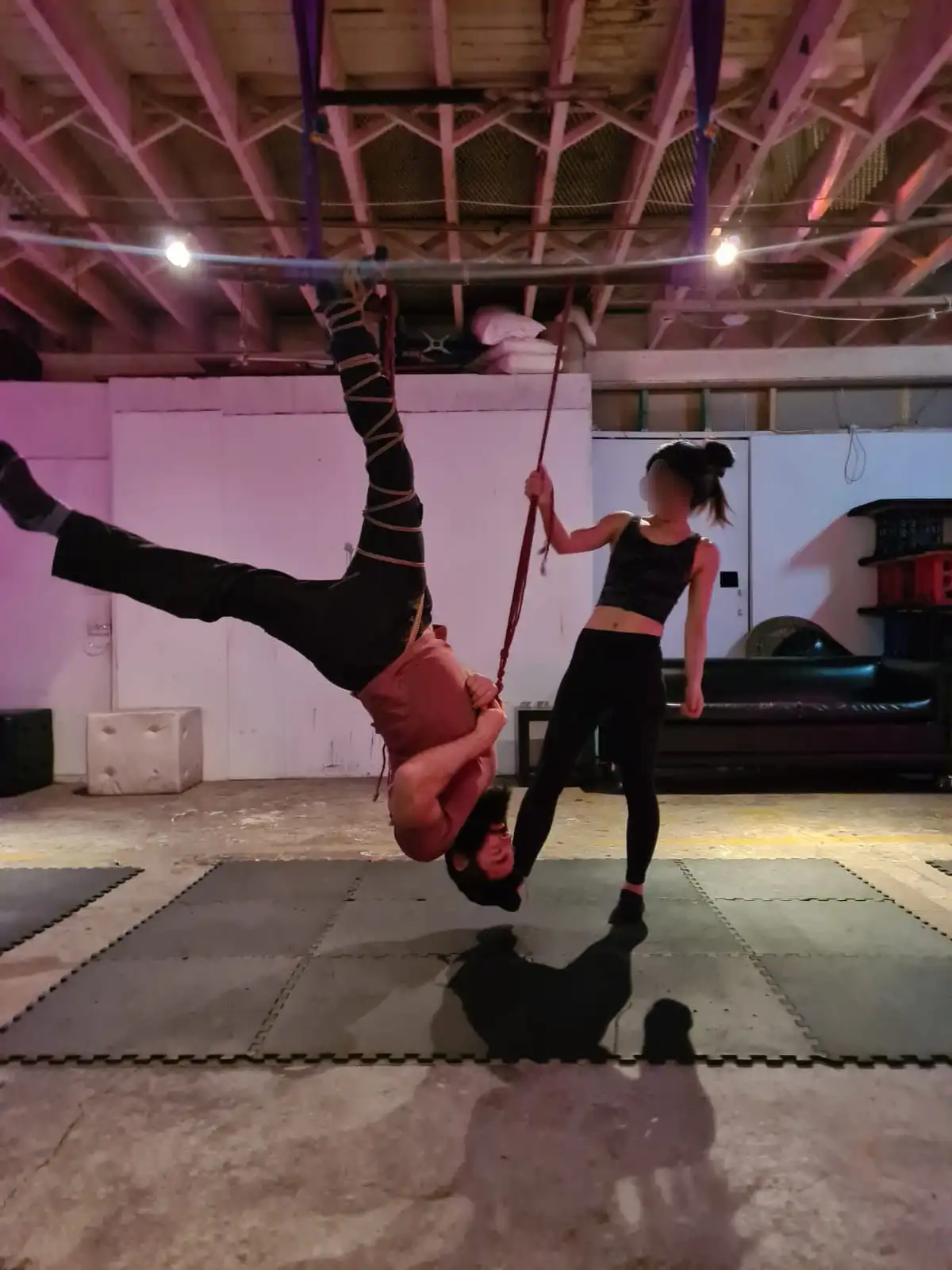A person is hanging upside down from a trapeze bar in a gym, their legs wrapped in straps, while another person stands…