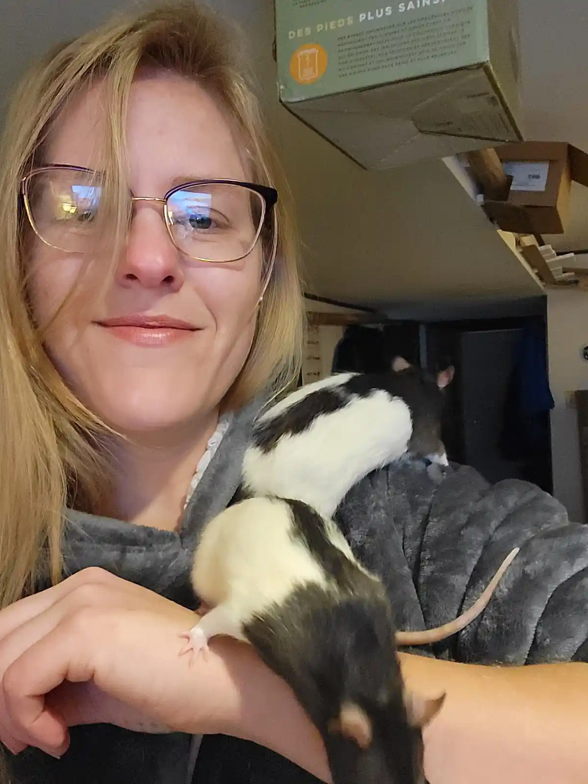 A woman with blonde hair and glasses smiles while holding two black-and-white rats, one on her shoulder and one on her…