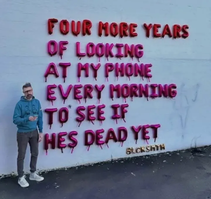 A man in a blue hoodie and glasses stands beside a white wall with large, dripping red and pink balloon letters spelling out…