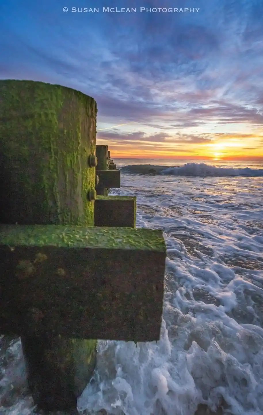 A serene sunset photograph shows waves crashing against moss-covered concrete breakwaters, with a vibrant sky of orange…