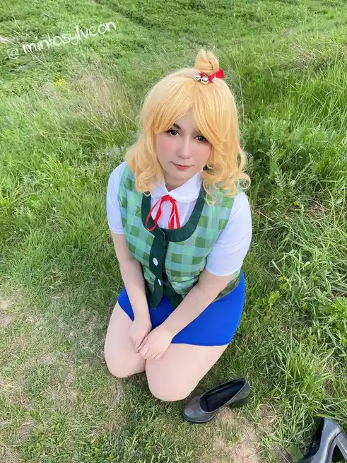 A person in a cosplay outfit with blonde hair tied in a bun with red and silver accessories, wearing a white shirt, green…